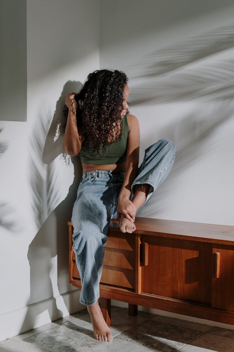 A woman with curly hair sits barefoot on furniture, bathed in tropical shadows.